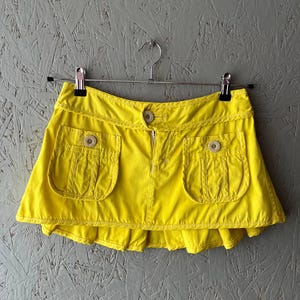May include: Bright yellow mini skirt with two front pockets and wooden buttons. The skirt has a ruffled hem and a zipper closure. The garment is hanging on a metal hanger against a gray background.