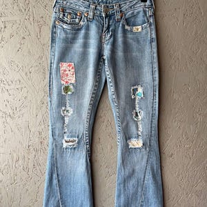 Puede incluir: Vaqueros de mezclilla azul claro con pernera acampanada, detalles desgastados y parches decorativos. Los vaqueros cuelgan de una percha de metal sobre un fondo neutro. Los parches presentan dise&ntilde;os florales y abstractos.