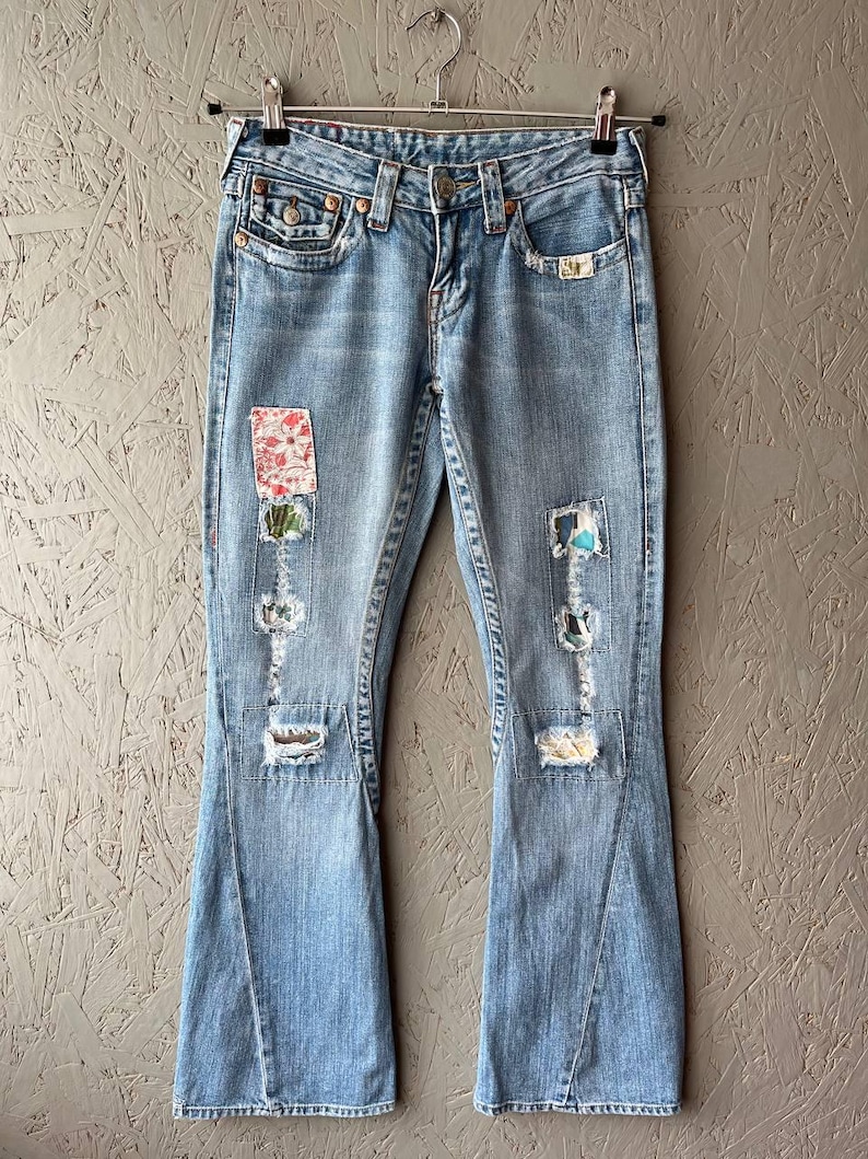 Puede incluir: Vaqueros de mezclilla azul claro con pernera acampanada, detalles desgastados y parches decorativos. Los vaqueros cuelgan de una percha de metal sobre un fondo neutro. Los parches presentan dise&ntilde;os florales y abstractos.