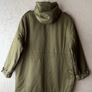 K&ouml;nnte beinhalten: Eine olivgr&uuml;ne Parka-Jacke mit Kapuze, langen &Auml;rmeln mit verstellbaren Schnallenb&uuml;ndchen, einer Taillenkordel und einer strukturierten Kapuze. Die Jacke ist von hinten abgebildet und zeigt eine glatte, einfarbige Stoffkonstruktion.