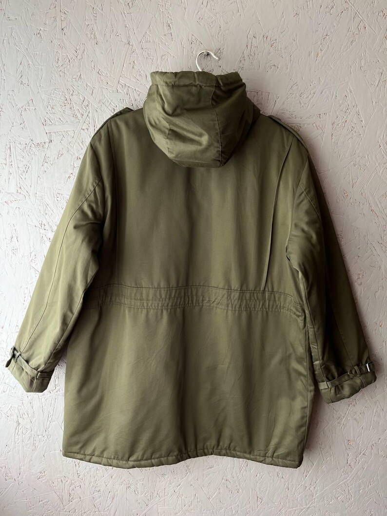 K&ouml;nnte beinhalten: Eine olivgr&uuml;ne Parka-Jacke mit Kapuze, langen &Auml;rmeln mit verstellbaren Schnallenb&uuml;ndchen, einer Taillenkordel und einer strukturierten Kapuze. Die Jacke ist von hinten abgebildet und zeigt eine glatte, einfarbige Stoffkonstruktion.