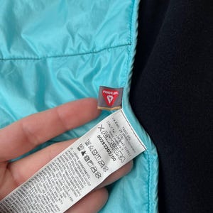 May include: A close-up shot of a turquoise quilted jacket section. The jacket features a black lining and a label with the "PRIMALOFT" brand and washing instructions.