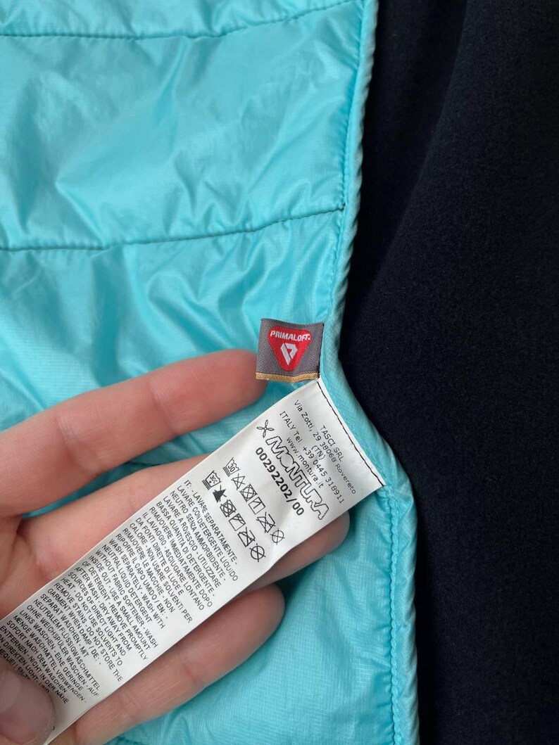 May include: A close-up shot of a turquoise quilted jacket section. The jacket features a black lining and a label with the "PRIMALOFT" brand and washing instructions.