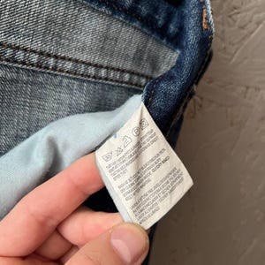 以下が含まれることがあります： 白いタグが付いたデニム衣料品のクローズアップ。タグには、記号とテキストによる洗濯表示が記載されています。デニムはミディアムブルーウォッシュで、ステッチが見え、内側はライトブルーです。