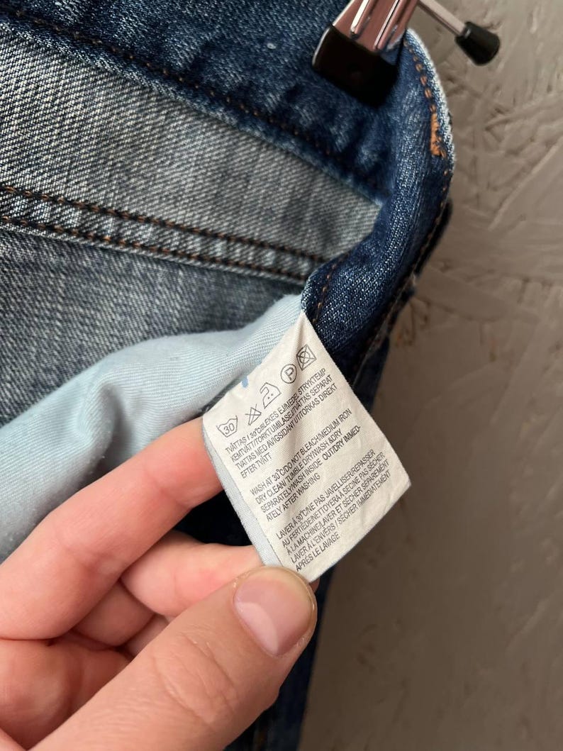 以下が含まれることがあります： 白いタグが付いたデニム衣料品のクローズアップ。タグには、記号とテキストによる洗濯表示が記載されています。デニムはミディアムブルーウォッシュで、ステッチが見え、内側はライトブルーです。