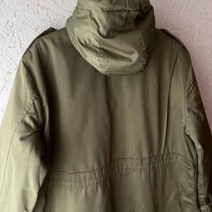 K&ouml;nnte beinhalten: Eine olivgr&uuml;ne Jacke mit Kapuze, die &uuml;ber eine strukturierte Kapuze, lange &Auml;rmel und eine horizontale Naht am R&uuml;cken verf&uuml;gt. Die Jacke ist aus glattem Stoff gefertigt und mit Schulterklappen versehen, was ein klares, funktionales Design ergibt.