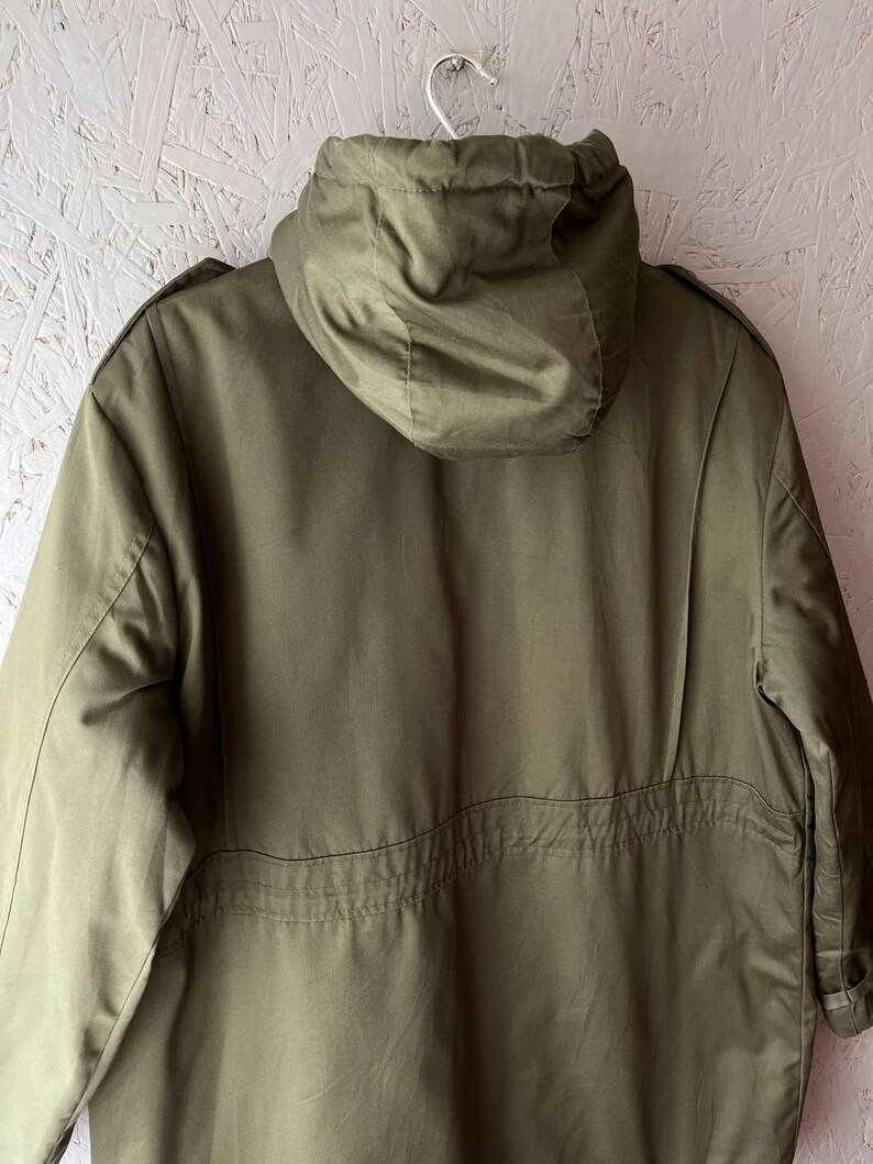 K&ouml;nnte beinhalten: Eine olivgr&uuml;ne Jacke mit Kapuze, die &uuml;ber eine strukturierte Kapuze, lange &Auml;rmel und eine horizontale Naht am R&uuml;cken verf&uuml;gt. Die Jacke ist aus glattem Stoff gefertigt und mit Schulterklappen versehen, was ein klares, funktionales Design ergibt.