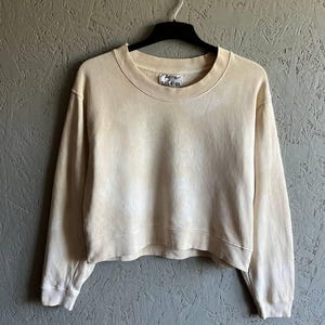 May include: A cream-colored cropped sweatshirt with a crew neck and long sleeves. The sweatshirt has a faded, tie-dye effect and features a small "Acne Studios" label on the front.