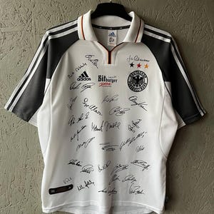 Könnte beinhalten: Weißes Adidas Deutschland Fußballtrikot mit schwarzen Akzenten auf den Ärmeln und Schultern. Das Trikot zeigt das Adidas-Logo, das deutsche Nationalmannschaftswappen und mehrere Unterschriften. Es hat einen Kragen und das Bitburger Drive-Logo.