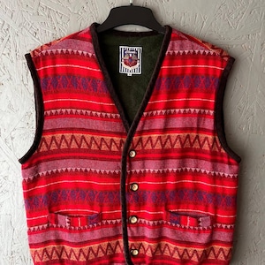 Op de afbeelding: Een rood vest met patroon met een V-hals en twee zakken. Het vest heeft een geometrisch ontwerp in tinten rood, paars en oranje. Het heeft een donkerbruine bies en houten knopen. Het vest hangt aan een zwarte hanger.
