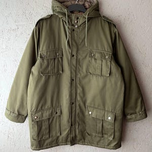 K&ouml;nnte beinhalten: Eine olivgr&uuml;ne Jacke mit Kapuze, Frontrei&szlig;verschluss und Druckknopfleiste, zwei Brusttaschen mit Klappen, zwei unteren Taschen mit Klappen, Schulterklappen sowie einem braunen, gesteppten Innenfutter, das in der Kapuze sichtbar ist.