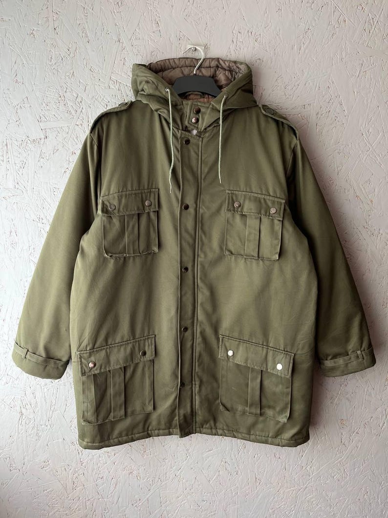 K&ouml;nnte beinhalten: Eine olivgr&uuml;ne Jacke mit Kapuze, Frontrei&szlig;verschluss und Druckknopfleiste, zwei Brusttaschen mit Klappen, zwei unteren Taschen mit Klappen, Schulterklappen sowie einem braunen, gesteppten Innenfutter, das in der Kapuze sichtbar ist.