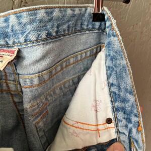 Puede incluir: Primer plano de unos vaqueros de mezclilla azul claro con costuras naranjas, colgados de una percha met&aacute;lica. El forro interior del bolsillo es blanco con un estampado rojo descolorido. Se ve una mano en la parte inferior derecha.