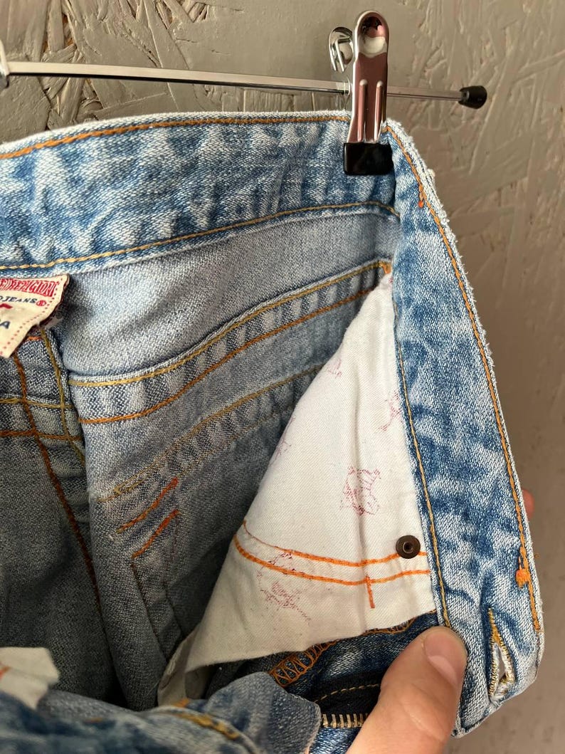 Puede incluir: Primer plano de unos vaqueros de mezclilla azul claro con costuras naranjas, colgados de una percha met&aacute;lica. El forro interior del bolsillo es blanco con un estampado rojo descolorido. Se ve una mano en la parte inferior derecha.