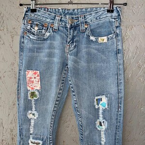 Puede incluir: Vaqueros de mezclilla azul claro con parches decorativos y detalles desgastados. Los vaqueros cuelgan de una percha met&aacute;lica. Los parches presentan patrones florales y abstractos, a&ntilde;adiendo un toque &uacute;nico al denim cl&aacute;sico.