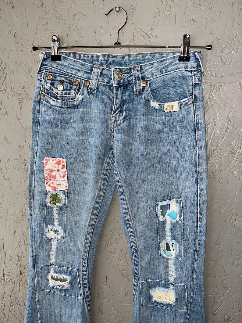 Puede incluir: Vaqueros de mezclilla azul claro con parches decorativos y detalles desgastados. Los vaqueros cuelgan de una percha met&aacute;lica. Los parches presentan patrones florales y abstractos, a&ntilde;adiendo un toque &uacute;nico al denim cl&aacute;sico.