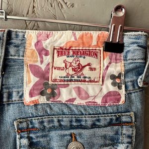 Puede incluir: Primer plano de unos vaqueros de mezclilla de lavado claro con un parche de la marca True Religion. El parche presenta un dise&ntilde;o floral en rosa, naranja y blanco, con el logotipo y el texto de la marca. Los vaqueros est&aacute;n sujetos por una pinza met&aacute;lica.