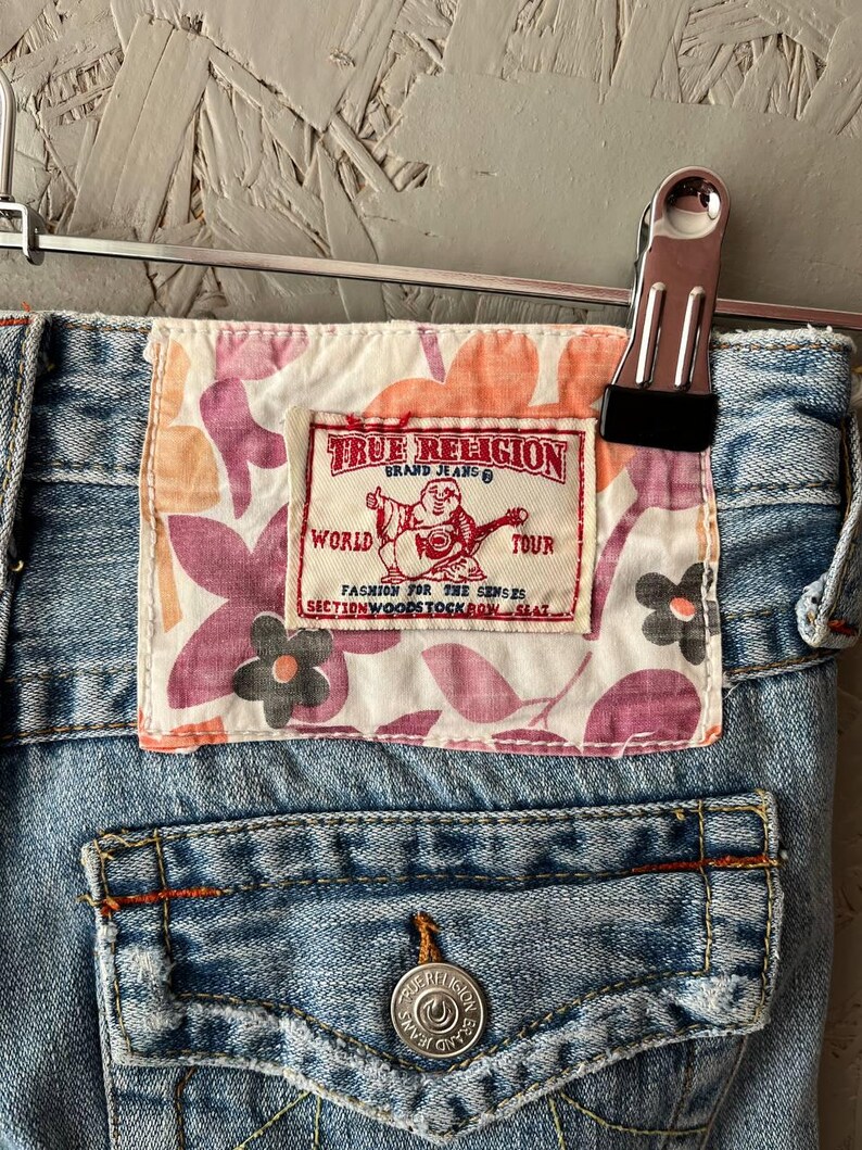 Puede incluir: Primer plano de unos vaqueros de mezclilla de lavado claro con un parche de la marca True Religion. El parche presenta un dise&ntilde;o floral en rosa, naranja y blanco, con el logotipo y el texto de la marca. Los vaqueros est&aacute;n sujetos por una pinza met&aacute;lica.