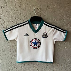 K&ouml;nnte beinhalten: Wei&szlig;es Fu&szlig;balltrikot mit schwarzen Streifen auf den Schultern und gr&uuml;nen Verzierungen. Das Adidas-Logo ist auf der Brust, zusammen mit einem Team-Emblem und einem runden Aufn&auml;her mit Text und einem blauen Stern.