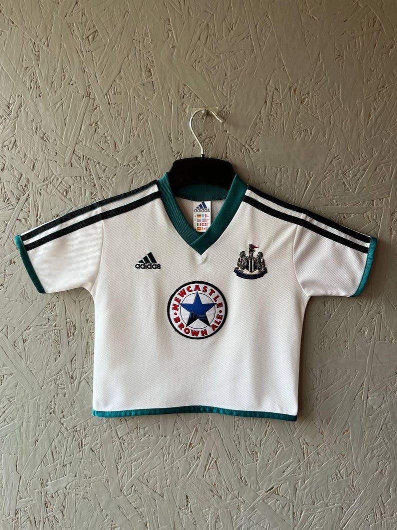 K&ouml;nnte beinhalten: Wei&szlig;es Fu&szlig;balltrikot mit schwarzen Streifen auf den Schultern und gr&uuml;nen Verzierungen. Das Adidas-Logo ist auf der Brust, zusammen mit einem Team-Emblem und einem runden Aufn&auml;her mit Text und einem blauen Stern.