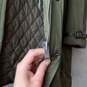 K&ouml;nnte beinhalten: Ein olivgr&uuml;ner Parka mit gestepptem Innenfutter, einem silberfarbenen Metallrei&szlig;verschluss mit YKK-Schieber und einer Druckknopfleiste. Der &Auml;rmel verf&uuml;gt &uuml;ber einen Schnallenriemen am B&uuml;ndchen und das Kleidungsst&uuml;ck ist aus strapazierf&auml;higem Stoff.