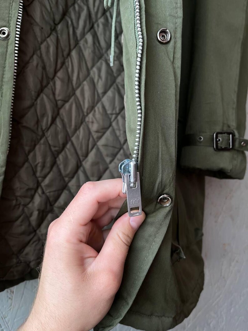 K&ouml;nnte beinhalten: Ein olivgr&uuml;ner Parka mit gestepptem Innenfutter, einem silberfarbenen Metallrei&szlig;verschluss mit YKK-Schieber und einer Druckknopfleiste. Der &Auml;rmel verf&uuml;gt &uuml;ber einen Schnallenriemen am B&uuml;ndchen und das Kleidungsst&uuml;ck ist aus strapazierf&auml;higem Stoff.