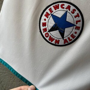 K&ouml;nnte beinhalten: Wei&szlig;es Sporttrikot mit t&uuml;rkisfarbenem Besatz und einem runden Aufn&auml;her. Der Aufn&auml;her zeigt einen blauen Stern, die Worte "NEWCASTLE BROWN ALE" in Rot und eine Schwarz-Wei&szlig;-Illustration.