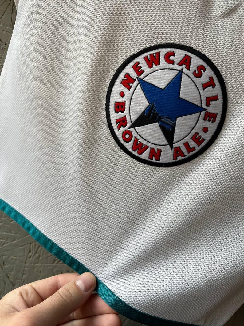 K&ouml;nnte beinhalten: Wei&szlig;es Sporttrikot mit t&uuml;rkisfarbenem Besatz und einem runden Aufn&auml;her. Der Aufn&auml;her zeigt einen blauen Stern, die Worte "NEWCASTLE BROWN ALE" in Rot und eine Schwarz-Wei&szlig;-Illustration.