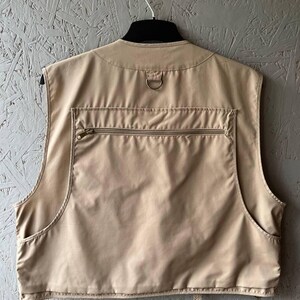 May include: A beige utility vest featuring a large horizontal zippered pocket across the back, a metal D-ring located at the nape of the neck, and two open side pockets. The garment is sleeveless with a high neckline and a structured back panel.