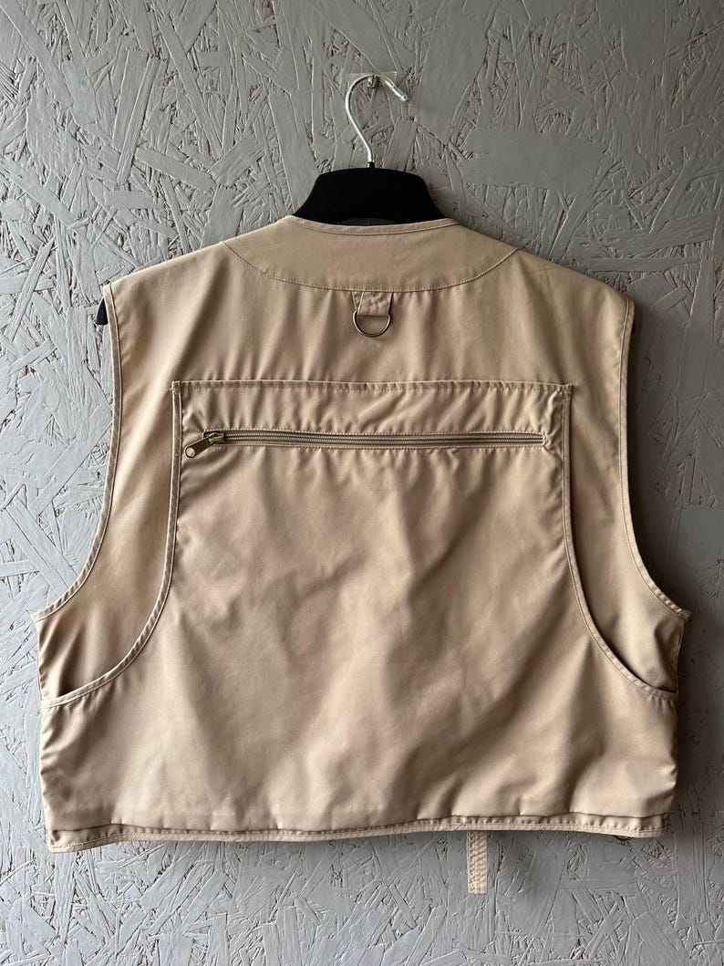 May include: A beige utility vest featuring a large horizontal zippered pocket across the back, a metal D-ring located at the nape of the neck, and two open side pockets. The garment is sleeveless with a high neckline and a structured back panel.