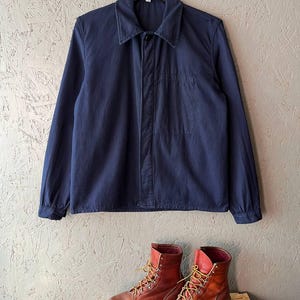 Könnte beinhalten: Eine marineblaue Arbeitsjacke mit einer einzelnen Brusttasche hängt über einem Paar brauner Leder-Arbeitsstiefel mit gelben Schnürsenkeln. Die Stiefel stehen auf einer Holzpalette.