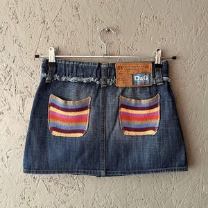 Puede incluir: Una falda vaquera con bolsillos a rayas coloridas y un parche de cuero con el texto "D&G" y "NEW VINTAGE". La falda está colgada en una percha de metal sobre un fondo gris texturizado.