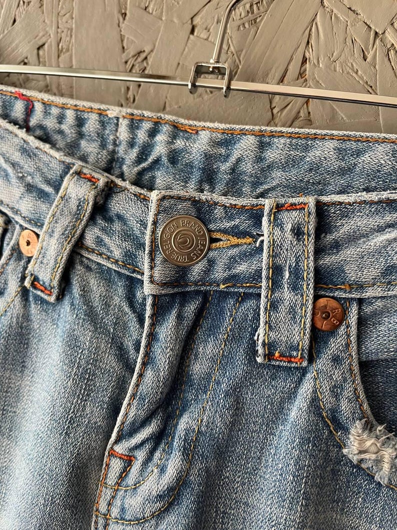 Puede incluir: Primer plano de unos vaqueros de mezclilla azul claro con costuras visibles y detalles de botones met&aacute;licos. Los vaqueros tienen un dise&ntilde;o cl&aacute;sico de cinco bolsillos con cierre de bot&oacute;n y cremallera. La mezclilla est&aacute; ligeramente desgastada.