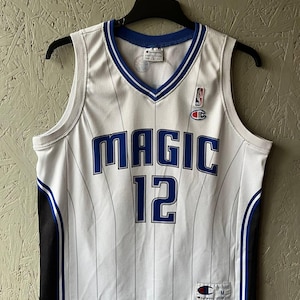 May include: White Champion basketball jersey with the word "MAGIC" and the number "12" in blue. The jersey has a blue and white V-neck collar and blue and black side panels. The NBA and Champion logos are visible.