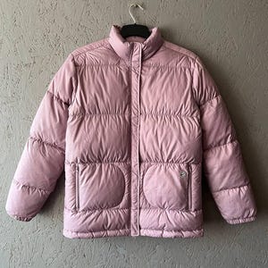 Puede incluir: Una chaqueta acolchada de color rosa claro con cierre de cremallera y dos bolsillos. La chaqueta tiene un diseño acolchado y un cuello alto.