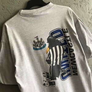 Puede incluir: Camiseta gris con un dibujo animado en blanco y negro de un &aacute;rbitro con una camiseta del Newcastle United. La camiseta tiene el escudo del equipo y las palabras "NEWCASTLE UNITED" en la espalda.