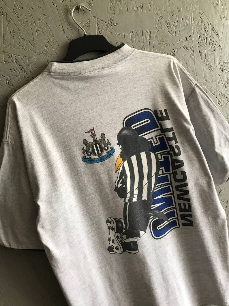 Puede incluir: Camiseta gris con un dibujo animado en blanco y negro de un &aacute;rbitro con una camiseta del Newcastle United. La camiseta tiene el escudo del equipo y las palabras "NEWCASTLE UNITED" en la espalda.