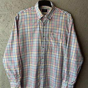 May include: A button-down shirt with a colorful plaid pattern in red, yellow, blue, and green on a white background. The long-sleeved shirt has a button-down collar and a small pocket on the chest. The shirt is hanging on a black hanger.