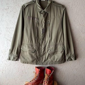 May include: An olive green field jacket with a collared neckline, shoulder epaulets, two vertical zippered chest pockets, and two lower flap pockets with snap closures, paired with a pair of brown leather lace-up work boots with yellow laces.