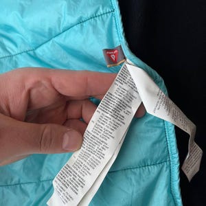 May include: A close-up shot of a turquoise quilted jacket's interior. The lining is a vibrant turquoise, contrasting with the black exterior. A white label with text provides details about the jacket's materials.