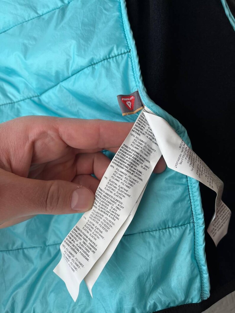 May include: A close-up shot of a turquoise quilted jacket's interior. The lining is a vibrant turquoise, contrasting with the black exterior. A white label with text provides details about the jacket's materials.
