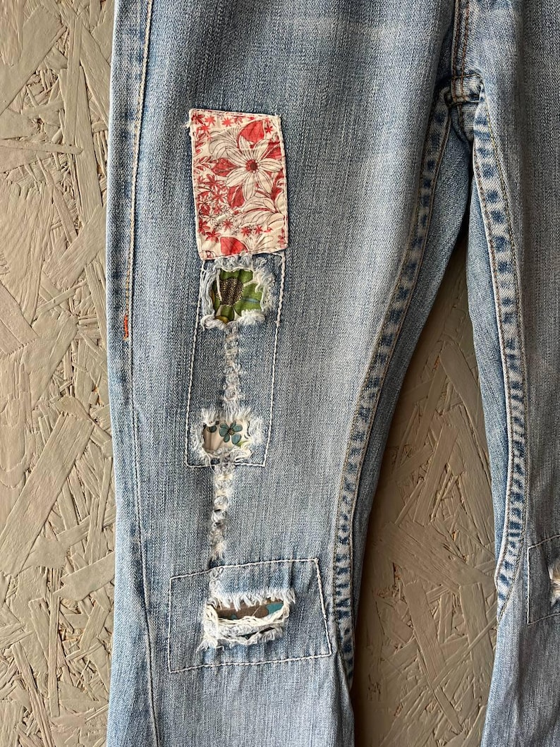 Puede incluir: Vaqueros de mezclilla azul claro con parches decorativos. Los parches son de tela floral en rojo y blanco, y patrones verdes y azules. Los vaqueros tienen detalles desgastados con costuras visibles.
