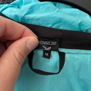 May include: A close-up of a turquoise jacket with a black trim. The jacket's tag displays the brand name "MONTURA" in white, along with a leaf logo and the size "M". The jacket is made of a lightweight, possibly water-resistant material.