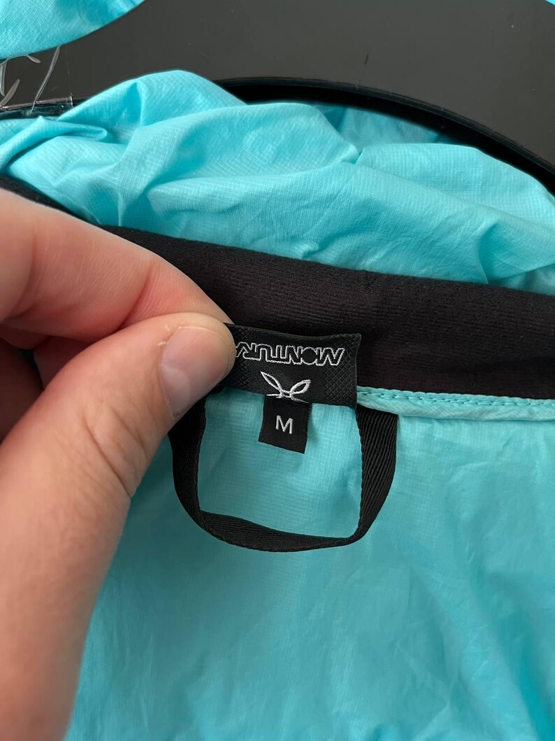 May include: A close-up of a turquoise jacket with a black trim. The jacket's tag displays the brand name "MONTURA" in white, along with a leaf logo and the size "M". The jacket is made of a lightweight, possibly water-resistant material.