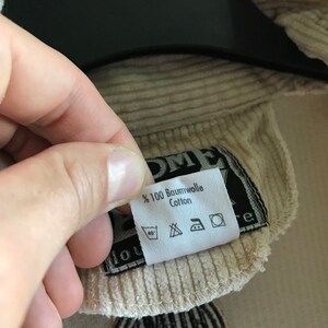 May include: A beige corduroy jacket with a black patch that says "100% Baumwolle Cotton" and has care instructions. The jacket has a black patch with the text "1 HUM 1 YOU" in white.