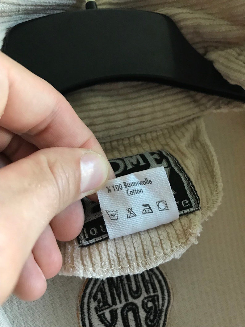 May include: A beige corduroy jacket with a black patch that says "100% Baumwolle Cotton" and has care instructions. The jacket has a black patch with the text "1 HUM 1 YOU" in white.