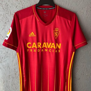 May include: A red soccer jersey with yellow accents, featuring the Adidas logo and the text "CARAVAN FRAGANCIAS". The jersey has a V-neck and a team crest on the chest. The jersey is hanging on a black hanger.