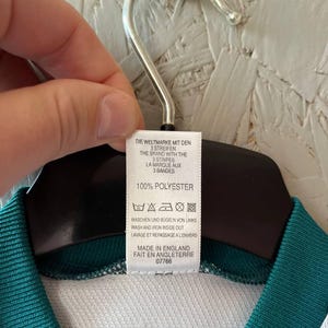 K&ouml;nnte beinhalten: Ein wei&szlig;es und tealfarbenes Sporttrikot mit einem Etikett mit der Aufschrift "DIE WELTMARKE MIT DEN 3 STREIFEN" und "MADE IN ENGLAND". Das Trikot besteht aus 100 % Polyester.