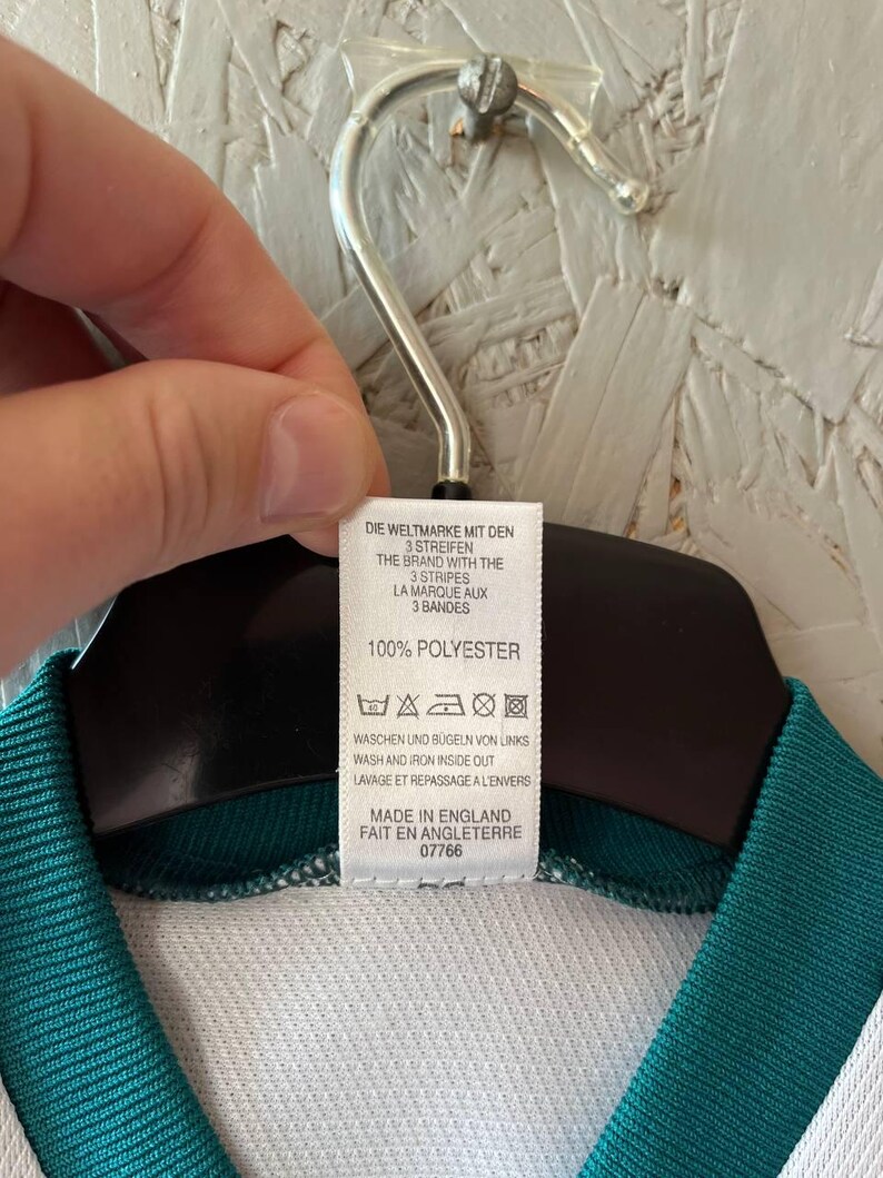 K&ouml;nnte beinhalten: Ein wei&szlig;es und tealfarbenes Sporttrikot mit einem Etikett mit der Aufschrift "DIE WELTMARKE MIT DEN 3 STREIFEN" und "MADE IN ENGLAND". Das Trikot besteht aus 100 % Polyester.