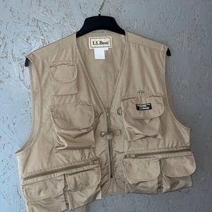 May include: A tan utility vest featuring multiple pockets, a front zipper closure, and an L.L.Bean brand label at the neckline. The garment includes a small embroidered name tag reading JESS and a rectangular L.L.Bean logo patch on the chest pocket.